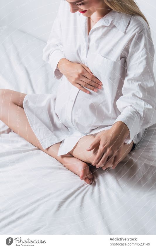 Anonyme schwangere Frau mit Hand auf dem Bauch auf dem Bett sitzend Angebot sanft mütterlich Harmonie positiv vorwegnehmen erwarten Schwangerschaft sorgenfrei