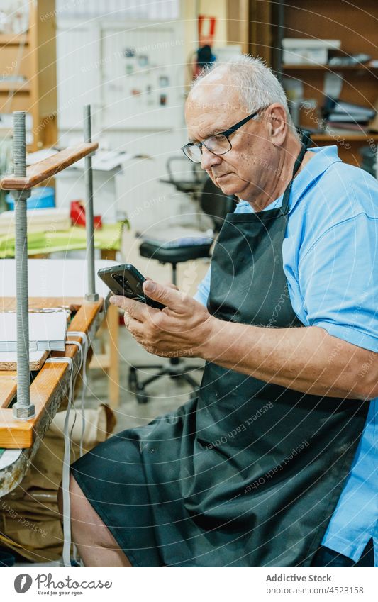 Fokussierter älterer männlicher Handwerker beim Surfen auf dem Smartphone in der Werkstatt Mann benutzend drucken Arbeit Konzentration lesen Hobelbank