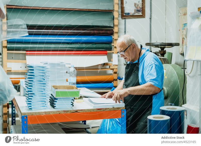 Aufmerksamer älterer Arbeiter bei der Papierkontrolle in einer Druckerei Mann prüfen drucken Kunstgewerbler achtsam Werkstatt Konzentration Papierkram Notebook