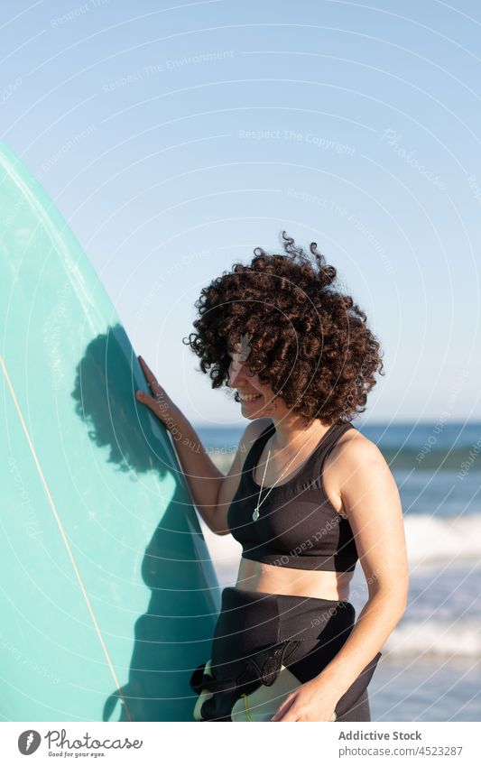 Lockenköpfige Frau mit Surfbrett am Meeresufer Surfer Lächeln MEER tropisch Strand winken schäumen Freude Glück sorgenfrei Aktivität Neoprenanzug froh Brandung