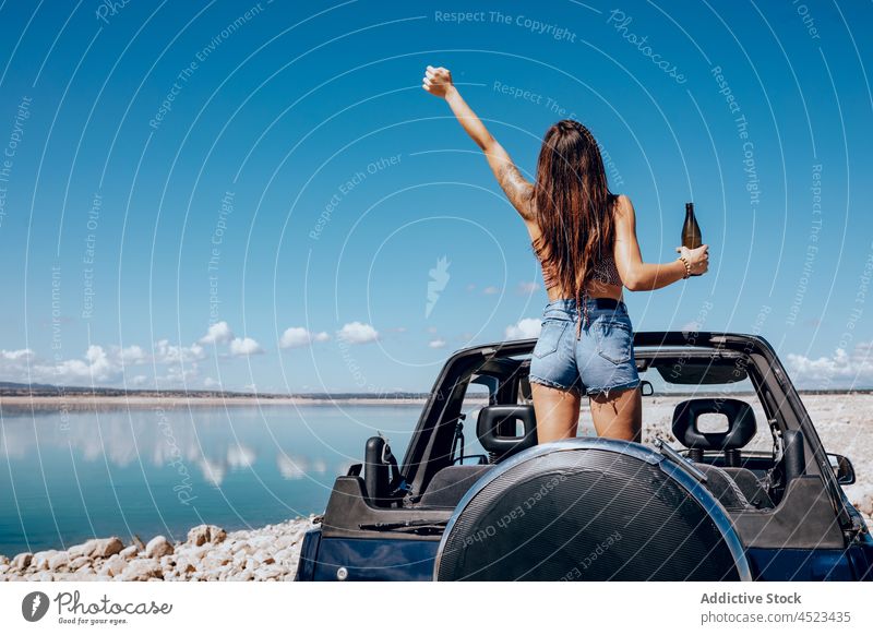 Fröhliche Frau auf Auto stehend mit erhobener Bierflasche PKW Safari Glück Flasche Fluss Kälte cool Freiheit Abenteuer sorgenfrei Arme hochgezogen
