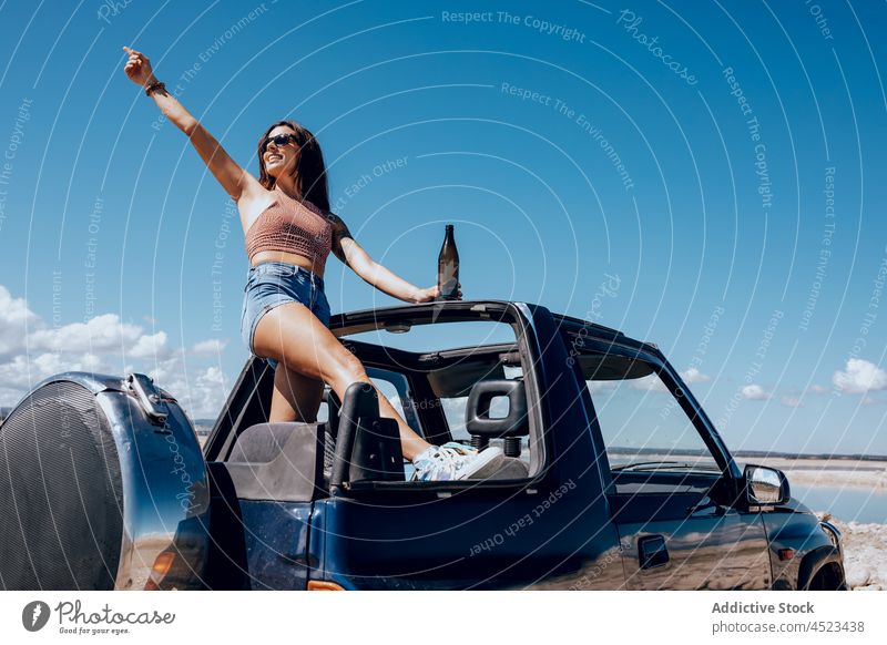Fröhliche Frau auf Auto stehend mit erhobener Bierflasche PKW Safari Glück Flasche Fluss Kälte cool Freiheit Abenteuer sorgenfrei Sonnenbrille