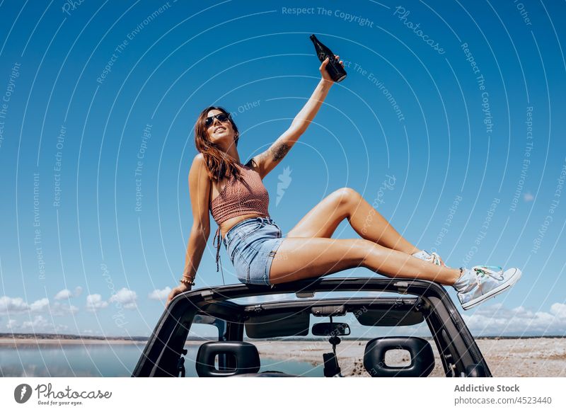 Fröhliche Frau auf Auto sitzend mit erhobener Bierflasche PKW Safari Glück Flasche Fluss Kälte cool Freiheit Abenteuer sorgenfrei Sonnenbrille