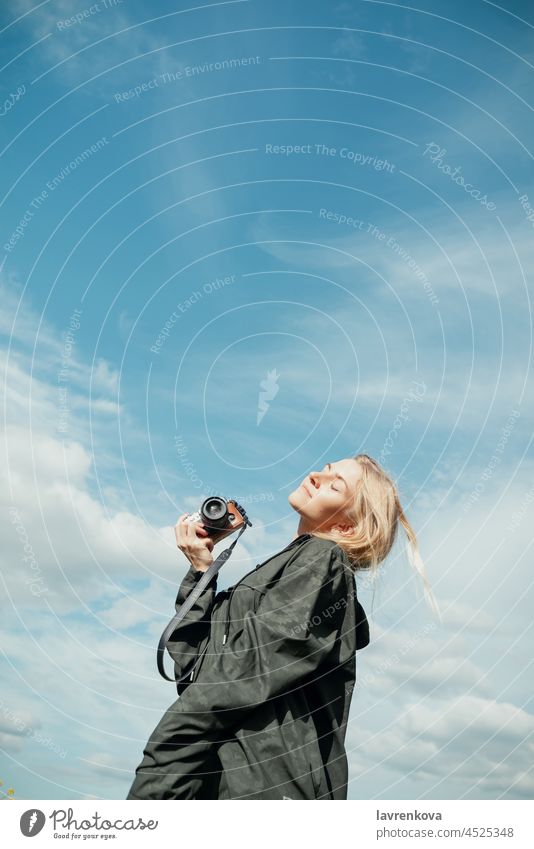 Junge blonde Frau in khakifarbenem Hemd mit Kapuze hält Kamera vor blauem Himmel im Freien Fotokamera Fotograf Augen geschlossen khakigrün hübsch Landschaft