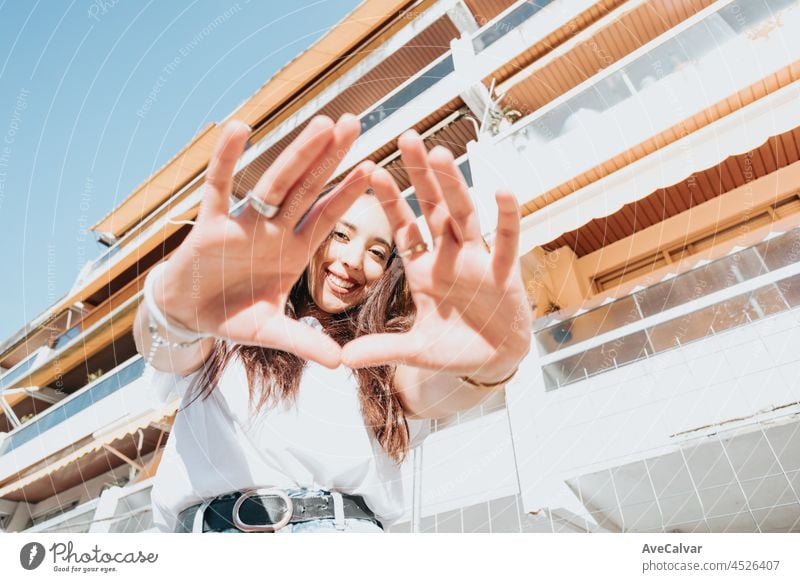 Close up Porträt der jungen arabischen Frau tun Zeichen tragen weißes Hemd und Blue Jeans lächelnd in die Kamera auf einem Block in der Kapuze. Straße Leben Stil, cool trendy. Soziales Netzwerk Konzept