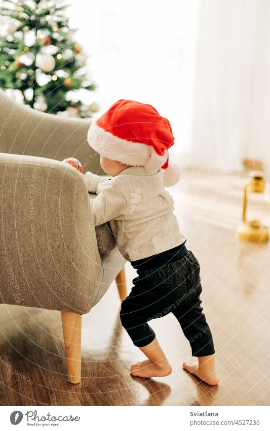 Ein kleiner Junge mit einer Weihnachtsmannmütze steht neben einem Sessel mit einem Weihnachtsbaumspielzeug in den Händen neben dem Weihnachtsbaum. Vorbereitung auf die Feiertage, festliche Dekoration