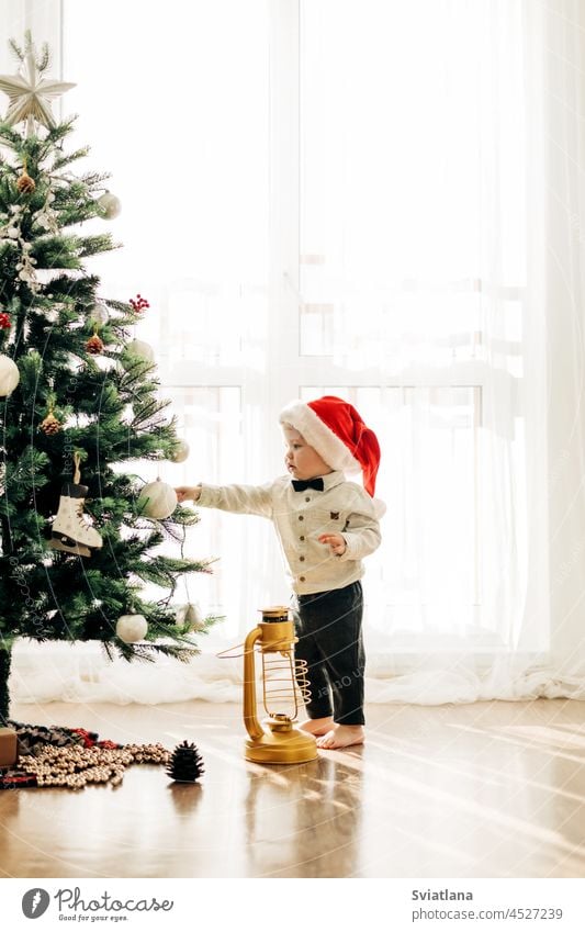 Ein kleines Kind mit einer Weihnachtsmütze vor dem Hintergrund eines Weihnachtsbaums. Weihnachten themed Bild, Postkarte. Baby Junge Kleinkind Winter