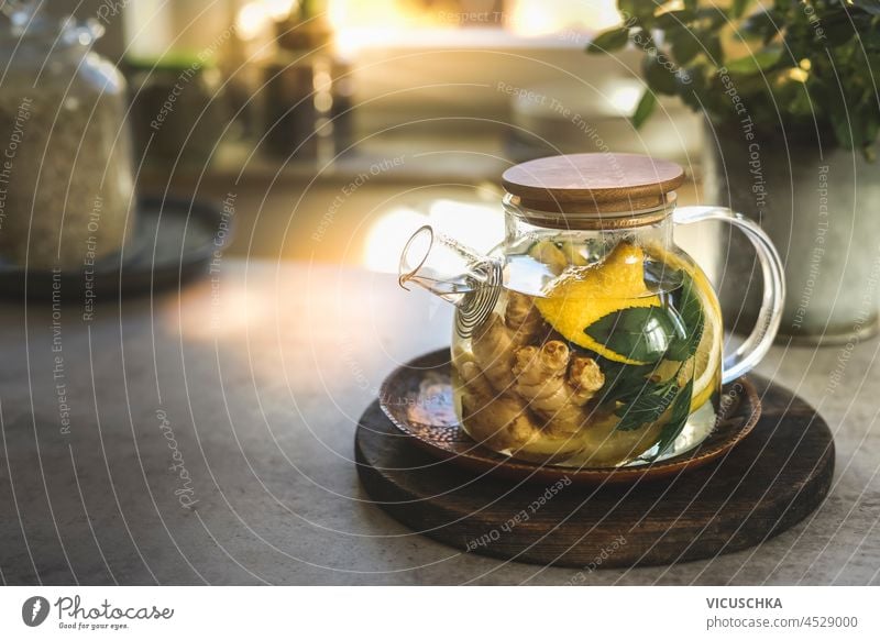 Glas-Teekanne mit Minzblättern, Zitrone und Ingwer auf Küchentisch mit Utensilien und Fenster Hintergrund. Kräutertee mit gesunden Zutaten und Vitamin C in der kalten Jahreszeit. Natürliche Behandlung. Vorderansicht.