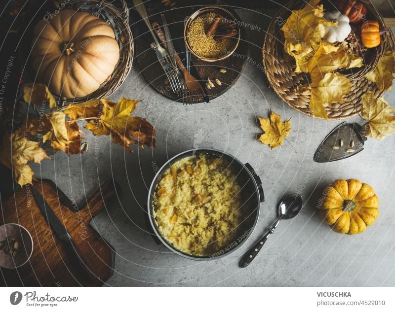 Hirsebrei mit Kürbissen im Kochtopf auf grauem Betontisch mit gelben Herbstblättern, Holzschneidebrett, Messer, Löffel und Küchenutensilien. Kochen gesundes veganes Frühstück zu Hause.Ansicht von oben