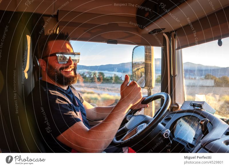 Ein Lkw-Fahrer, der froh ist, einen Lkw fahren zu können, und mit dem Daumen nach oben die Geste "OK" macht. Lastwagen Lenkrad arbeiten Glück Sonnenuntergang