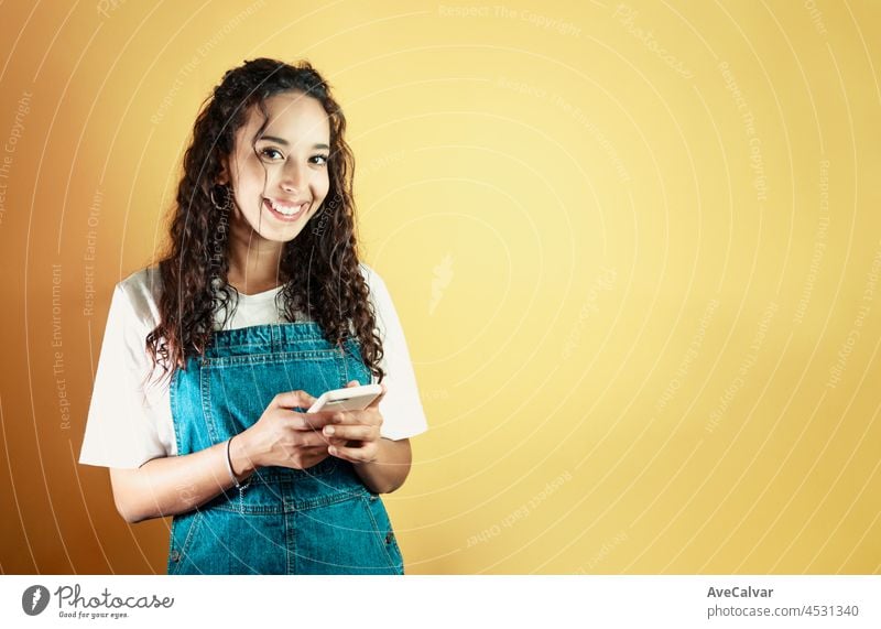 Junge gemischtrassige Frau, die ein Telefon hält, eine Nachricht schreibt und in die Kamera lächelt, gelber Hintergrund, abnehmbarer Hintergrund, minimales Bild, Kopierraum, grundlegende Situationen und Gefühlsausdrücke