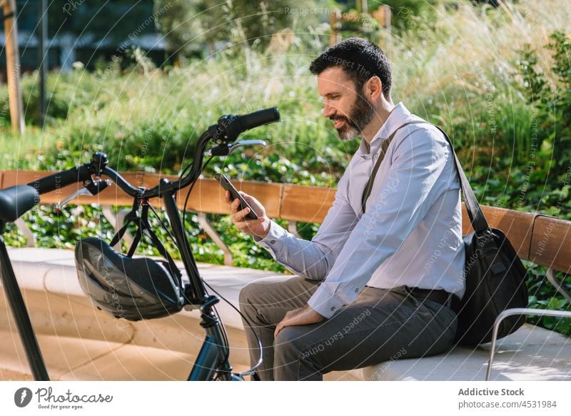 Ernster Mann, der auf einer Bank im Park auf sein Smartphone schaut Manager zuschauen benutzend lesen prüfen Nachricht Fahrrad männlich Apparatur Browsen