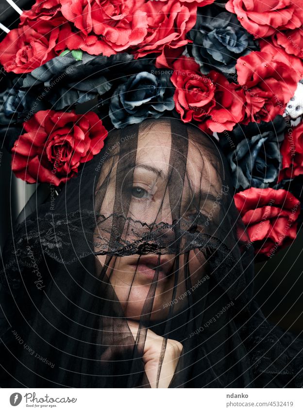 eine Frau kaukasischen Aussehens ist mit einem schwarzen Schleier und einem großen Kranz aus rot-schwarzen Rosen bekleidet Imkerschleier Totenkranz Schönheit