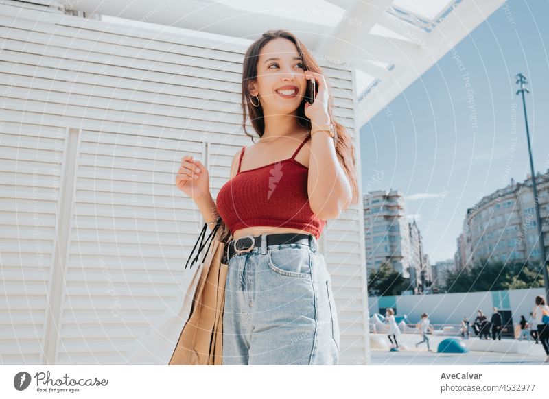 Junge arabische Frau, die einen Anruf tätigt, während sie Einkaufstaschen hält, Einkaufstag, modernes Outfit-Styling, lächelnd an einem sonnigen Tag kaufen