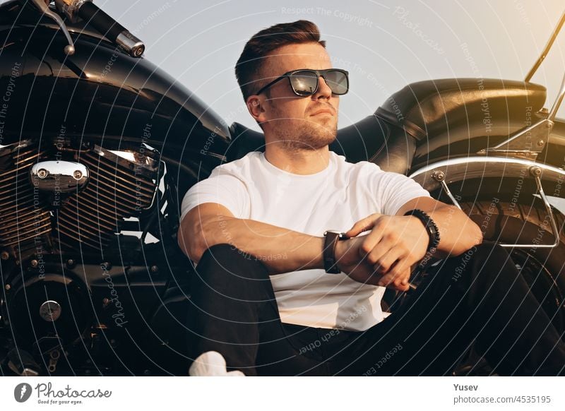Junger attraktiver Mann kaukasischer Ethnizität sitzt neben einem Motorrad. Netter süßer Mann mit Sonnenbrille, weißem T-Shirt und schwarzen Jeans. Lebensstil Fotografie.
