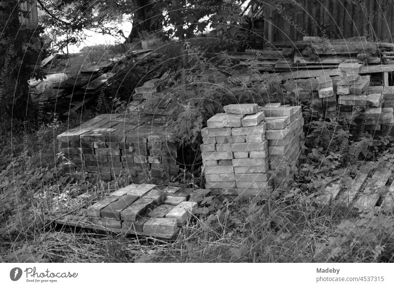 Sorgfältig gestapelte alte Bausteine im Schatten alter Bäume vor einer Holzhütte auf einem Bauernhof in Rudersau bei Rottenbuch im Kreis Weilheim-Schongau in Oberbayern, fotografiert in neorealistischem Schwarzweiß