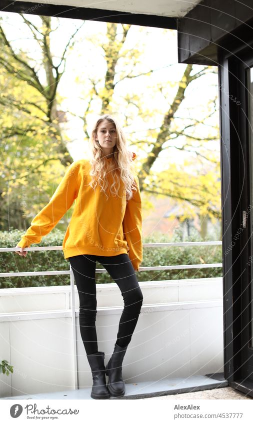 Ganzkörperportrait einer jungen, schlanken Frau mit langen, blonden Haaren und gelben Kapuzenpullover auf dem Balkon im Herbst junge Frau groß langhaarig schön