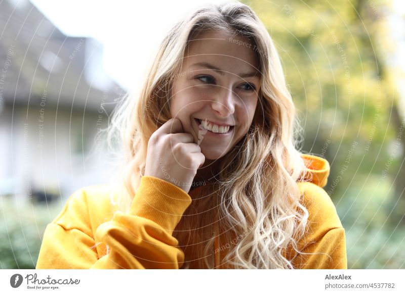 Portrait einer jungen, schlanken lächelnden Frau mit langen, blonden Haaren und gelben Kapuzenpullover auf dem Balkon im Herbst junge Frau groß langhaarig schön