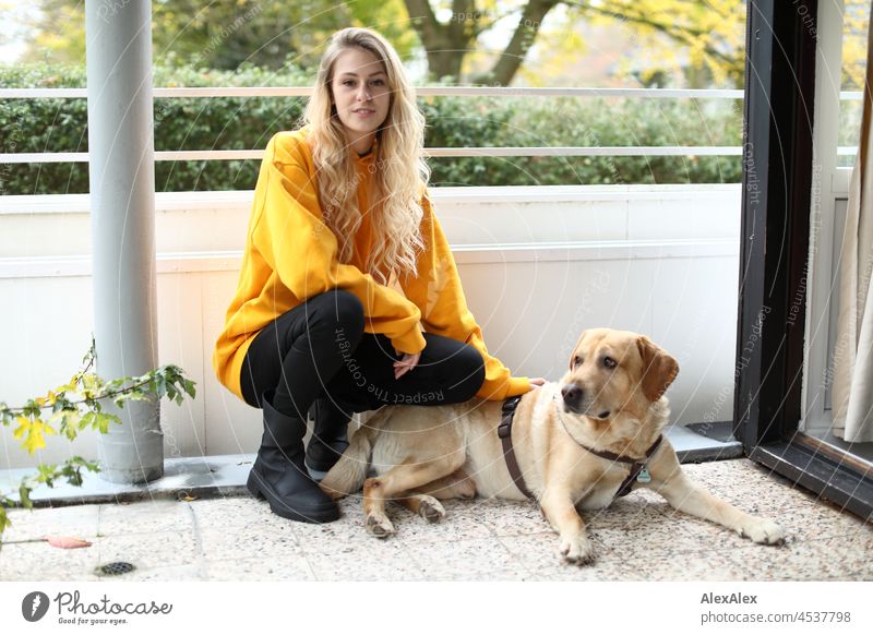 Junge, große, blonde, schlanke Frau hockt neben einem blonden Labrador auf dem Balkon junge Frau langhaarig schön anmutig athletisch ästhetisch sportlich
