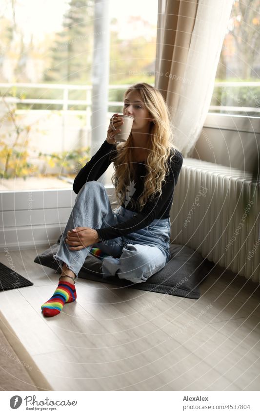 Junge, schlanke, große Frau mit langen, blonden Haaren sitzt im Wohnzimmer vor dem Balkonfenster und trinkt aus einer großen Tasse Tee oder Kaffee junge Frau