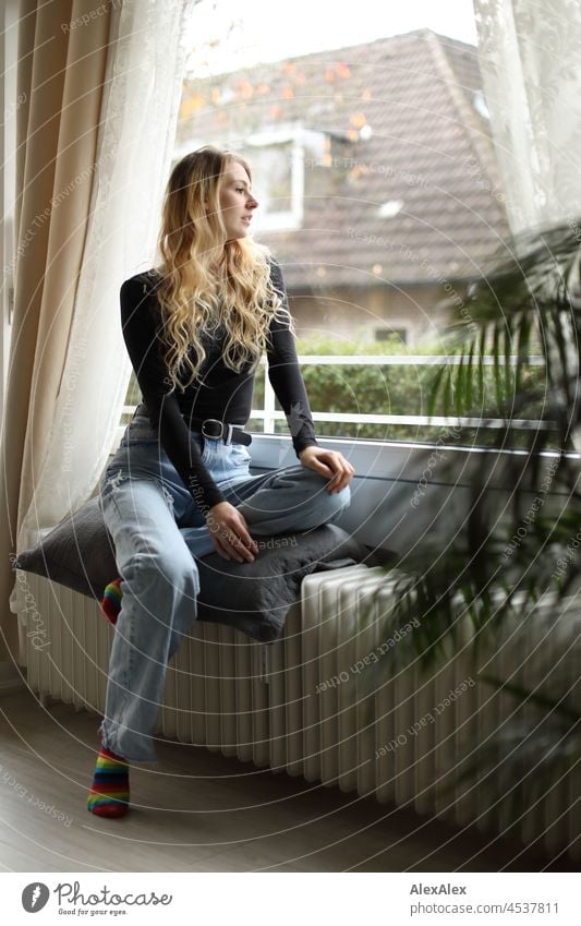Junge, schlanke, große Frau mit langen, blonden Haaren sitzt im Wohnzimmer vor dem Balkonfenster auf der Heizung junge Frau langhaarig schön anmutig Socken