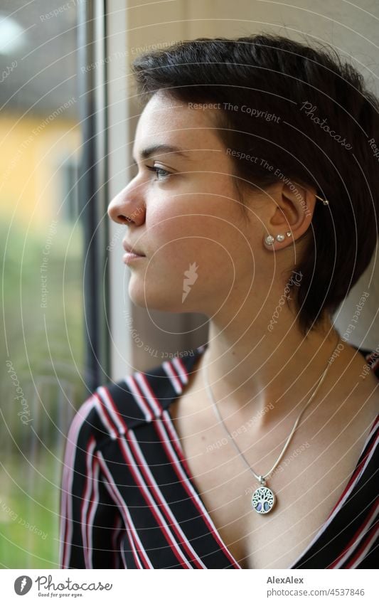Junge Frau mit kurzen, brünetten Haaren schaut aus dem Fenster junge Frau kurze Haare hübsch schauen natürlich authentisch Schmuck Kette Ohrringe schlank
