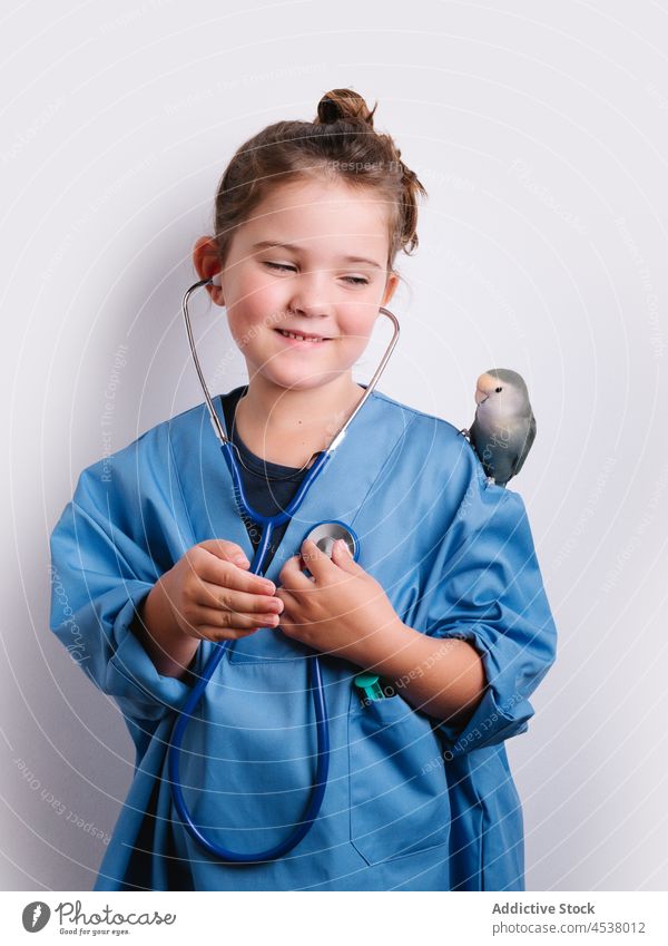Lächelndes Mädchen in Arztkleidung mit Papagei Stethoskop Uniform Verkleidung spielen medizinisch spielerisch Kindheit Sanitäter Instrument Vogel Herzschlag