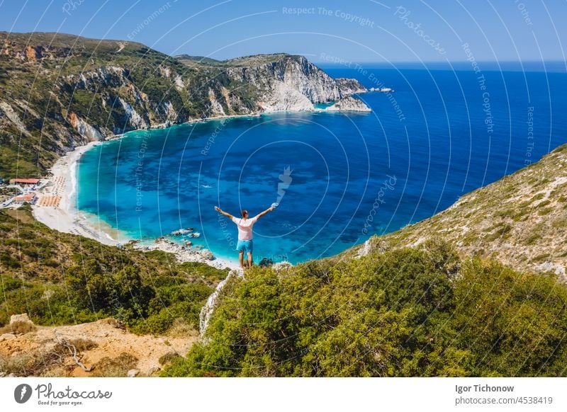 Mann, der auf einem Felsen steht und mit einem aufregenden Gefühl der Freiheit die Hände hebt, mit Blick auf den Strand von Petani. Insel Cephalonia, Griechenland
