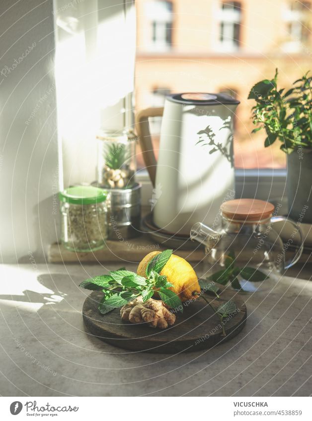 Zubereitung von frischem Ingwertee mit gesunden Zutaten zu Hause. Ingwer, Minzblätter und Zitrone auf Küchentisch mit Glas Teekanne, Wasserkocher und Gläser am Fenster Hintergrund.  Vorderansicht.