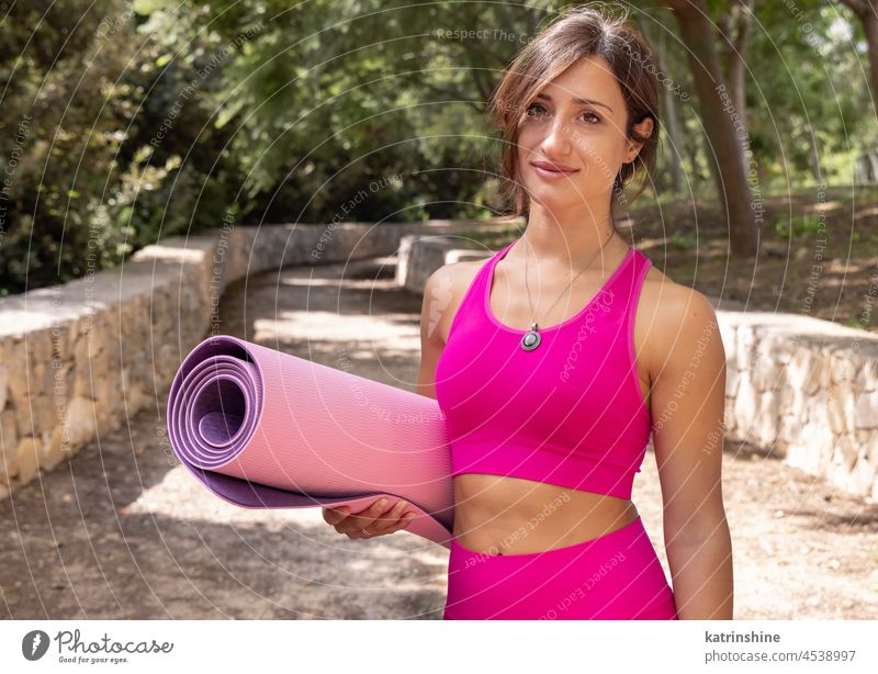 Lächelnde Frau in Sportkleidung mit gerollter Pilates-Matte in einem Park Sportbekleidung Aufenthalt Attrappe Unterlage Glück allein Gesundheit Sommer rosa grün