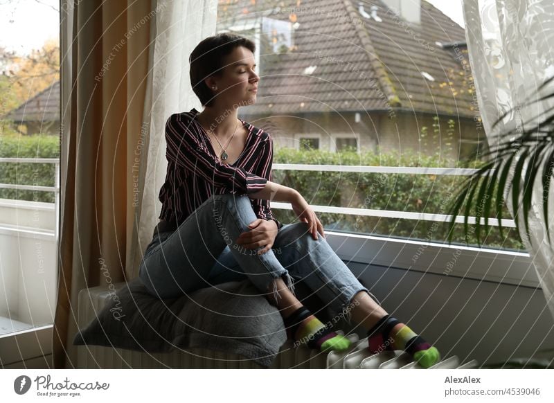 Junge Frau mit kurzen, brünetten Haaren sitzt auf der Heizung vorm Fenster im Wohnzimmer junge Frau kurze Haare hübsch schauen natürlich authentisch Schmuck