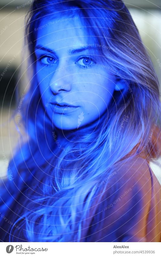 Portrait einer jungen, schlanken Frau mit langen, blonden Haaren im Wohnzimmer vor dem Fenster, sie ist mit blauem Licht angestrahlt junge Frau groß langhaarig