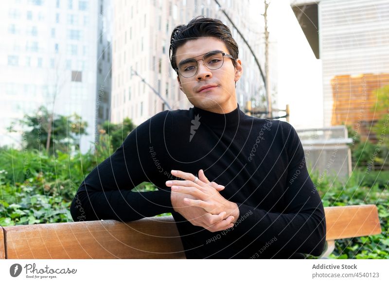 Junger asiatischer Mann in der Stadt Gebäude Straße Großstadt Stil urban modern sitzen Bank Outfit Vorschein ethnisch Bekleidung trendy Glück froh Typ Lifestyle