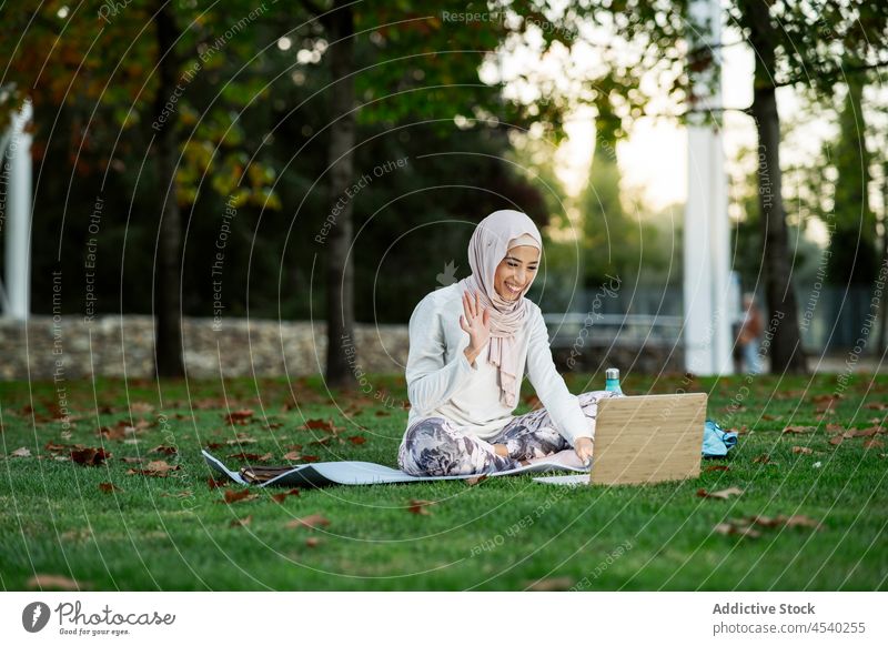Positive arabische Frau mit Videoanruf im Park Laptop Video-Chat Wellenhand online Kommunizieren Freizeit Hijab Rasen Browsen Zeitvertreib Netbook Gruß