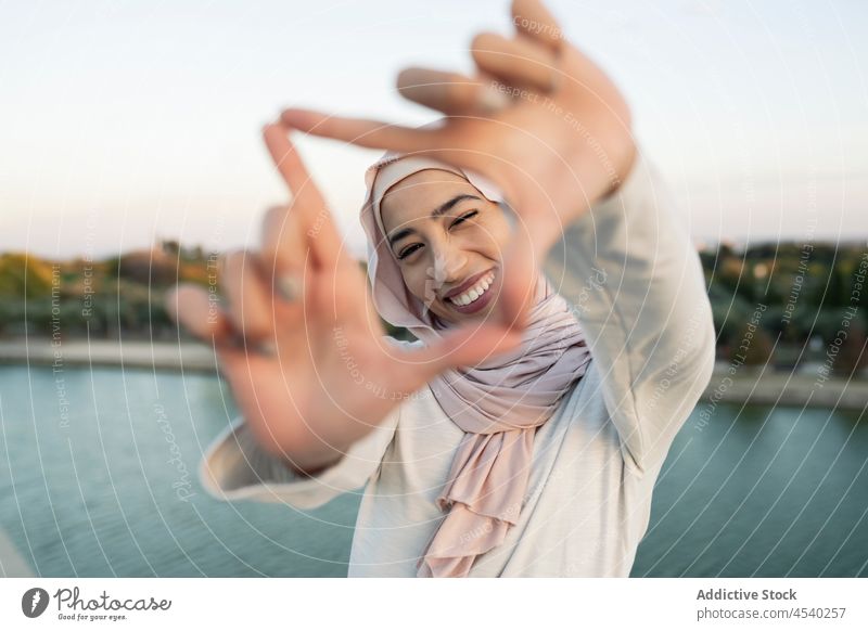 Fröhliche arabische Frau mit Rahmengeste auf dem Damm Hafengebiet Stauanlage Hijab Fluss Wasser Freizeit Ufer gestikulieren Zeitvertreib Flussufer muslimisch