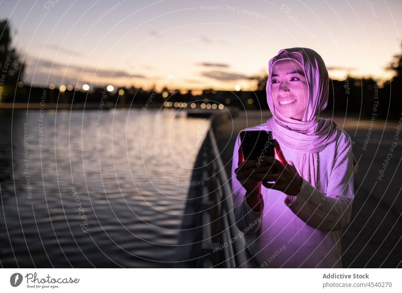 Positiv eingestellte muslimische Frau, die ihr Smartphone an einer Böschung benutzt benutzend purpur Licht Sonnenuntergang positiv Stauanlage dunkel Anschluss