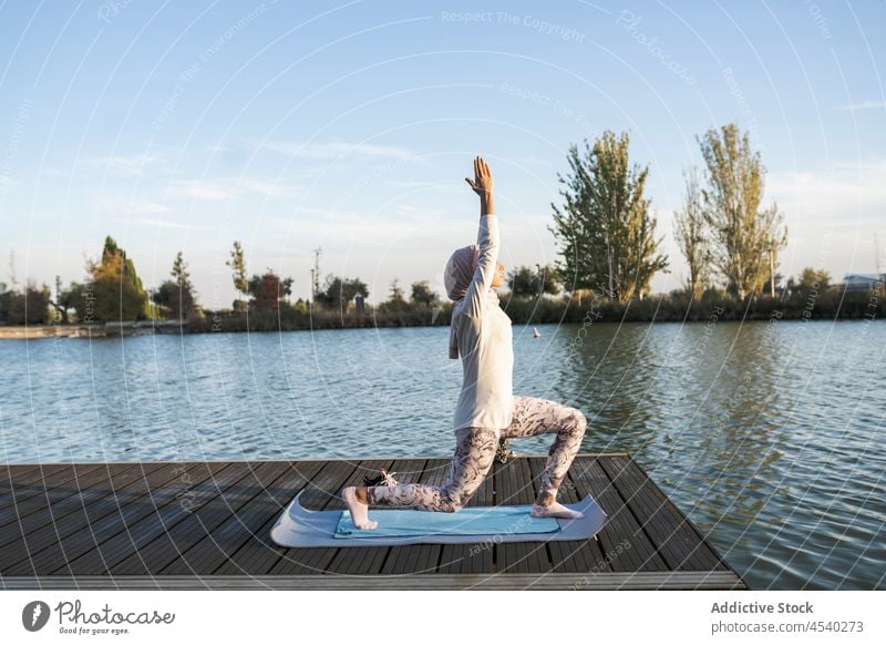 Anmutige arabische Frau in Anjaneyasana-Pose auf dem Pier meditieren anjaneyasana üben Yoga Körperhaltung Gleichgewicht Gelassenheit Asana Harmonie Hijab