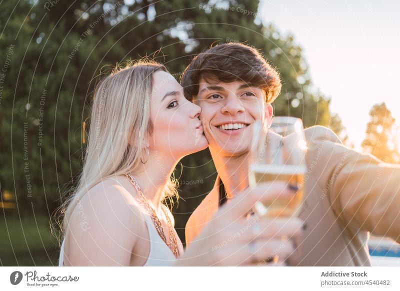 Frau mit Glas küsst Partner für gemeinsames Selfie im Park Paar heiter Kuss Alkohol Zusammensein Smartphone Funktelefon Champagne Selbstportrait sorgenfrei