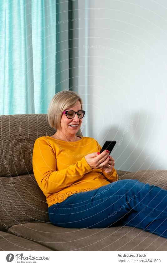 Lächelnde reife Frau mit Smartphone auf bequemem Sofa benutzend Internet digital Anschluss positiv Kälte Zeitvertreib Nachricht Mobile Apparatur online Brille