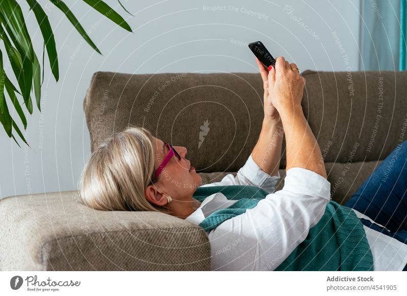 Frau mit Brille liegt auf der Couch und surft auf dem Smartphone benutzend sorgenfrei Kälte digital zuschauen Internet online positiv Gerät Mobile Lebensmitte