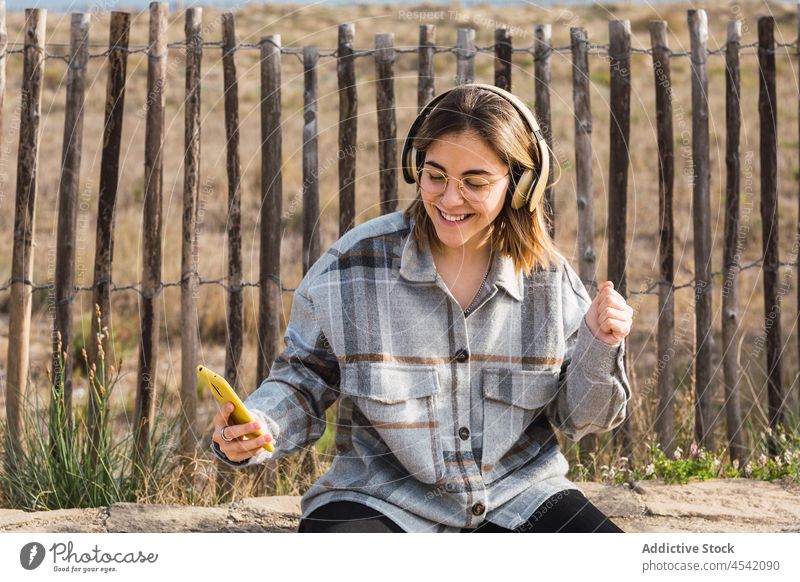 Glückliche junge Frau, die ein Smartphone benutzt und mit Kopfhörern in der Natur Musik hört zuhören Lächeln Landschaft Wiedergabeliste meloman Nachricht