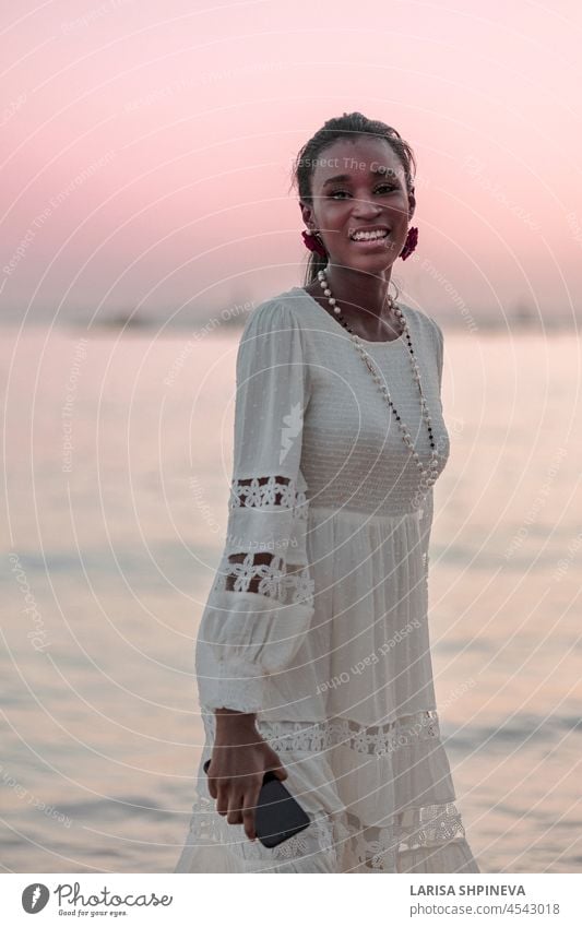 Schöne afrikanische Frau in Sonnenhut geht am Strand bei Sonnenuntergang. Mädchen Blogger hält Telefon in der Hand am Meer. Afrikanisch MEER schön schwarz