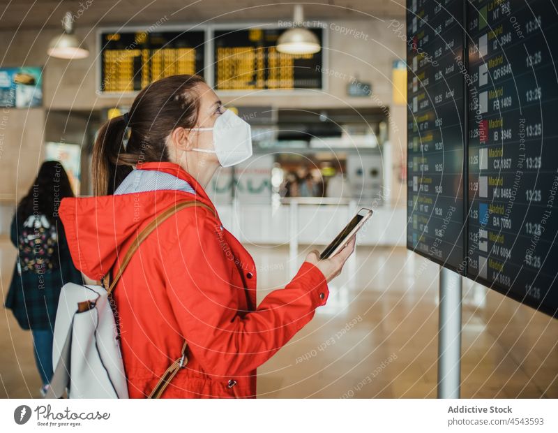 Frau mit medizinischer Maske surft auf einem Mobiltelefon in der Nähe eines Flugplans im Flughafen Reisender Ausflug Terminal Abheben Zeitplanung Schild