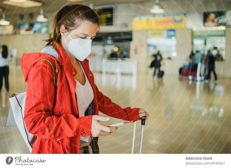 Frau mit medizinischer Maske surft im Flughafen auf ihrem Smartphone Browsen online Reisender Ausflug Abheben Mundschutz Koffer neue Normale schützend