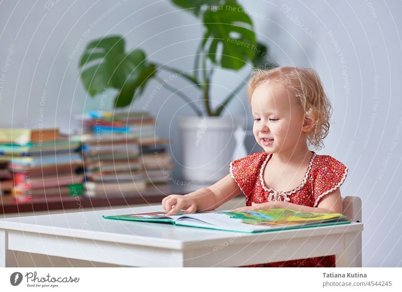 Ein süßes Mädchen liest ein Kinderbuch und hält einen Finger. Baby lesen Buch Lernen heimwärts Kleinkind Vorschulkind lockig Glück wenig jung Bildung studierend
