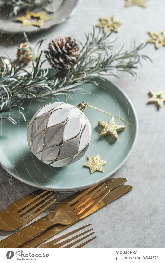 Weihnachtlich gedeckter Tisch mit weißer Vintage-Kugel, goldenen Deko-Sternen, Tannenzapfen, goldenem Besteck, tannengrünem und hellblauem Teller auf grauem Beton-Küchentisch. Ansicht von oben.