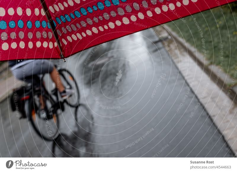 Ein Radfahrer fährt im Regen an einem Spaziergänger mit einem gepunkteten Regenschirm vorbei. radfahrer rot Straße Spiegelung gepunkteter Regenschirm
