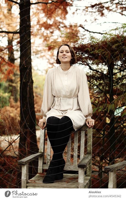 Junge Frau sitzt auf einer Holzstuhllehne im Park im Herbst und lächelt in die Kamera heiter Leichtigkeit beige grün schönes Wetter Stimmung Frauengesicht