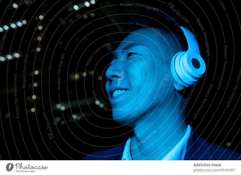 Fröhlicher asiatischer Mann mit Kopfhörern auf dunkler Straße Musik zuhören Nacht Gesang meloman formal Zeitvertreib Melodie Gerät Audio dunkel Dunkelheit
