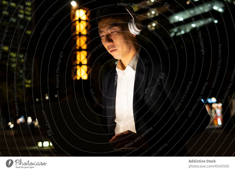 Asiatischer Geschäftsmann mit Laptop und Kopfhörern auf einer dunklen Straße Mann Gebäude Nacht Unternehmer Arbeit Funktelefon abgelegen Anzug Outfit elegant